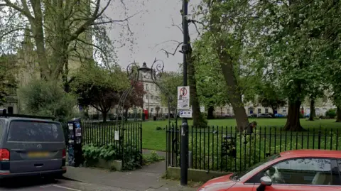 Portland Square, a small grassy park surrounded by black mteal fencing. There are several trees in the park, and a red car and grey van are parked on the road outside it.