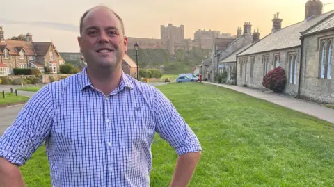 Guy Renner-Thompson is a man in his 30s wearing a blue short-sleeved shirt with a receding hairline. He is standing in a traditional English village and behind him is Bamburgh Castle an imposing large Victorian building on an outcrop sitting above the village.