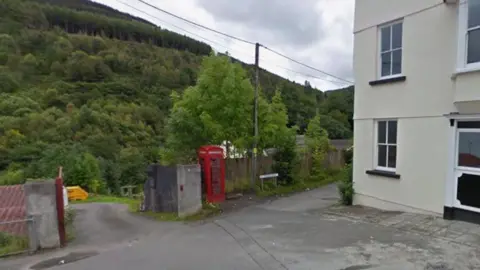 Google A red phone box next to a cream coloured building with green tree-lined mountains in the background. 
