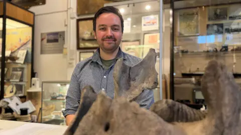 Dr Neil Adams, stands inside Holyhead museum. He’s smiling and facing the camera, wearing a checked shirt. In the foreground, slightly out of focus, is a large fossilised jawbone of a woolly mammoth, placed on a table.