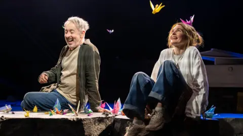 Tommy Ga-Ken Wan Two performers sit on the edge of a stage set, surrounded by brightly coloured origami cranes scattered around and suspended in the air. Dramatic spotlights illuminate the scene against a dark background, drawing attention to the vivid paper sculptures and casual costumes. The theatrical setting creates a striking contrast between the simple staging and the whimsical, dreamlike display of floating paper birds.