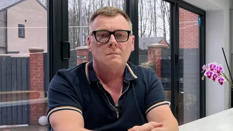 Graeme Blenkiron, a man with glasses and a navy polo shirt, is sitting at a table next to some cut flowers and an orchid.