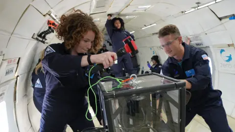 Steve Boxall Two young researchers in blue jump suits work on some equipment on a low-gravity flight. 