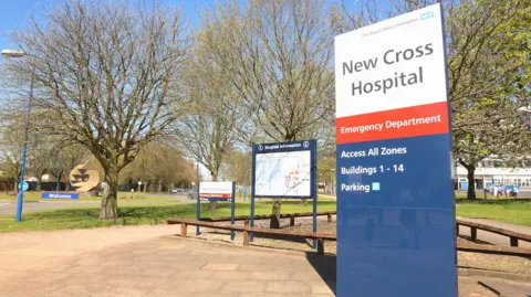 Royal Wolverhampton and Walsall Healthcare NHS Trusts Outside entrance of New Cross Hospital, trees in the back ground with a sign with NEW CROSS HOSPITAL on it with writing saying emergency department below