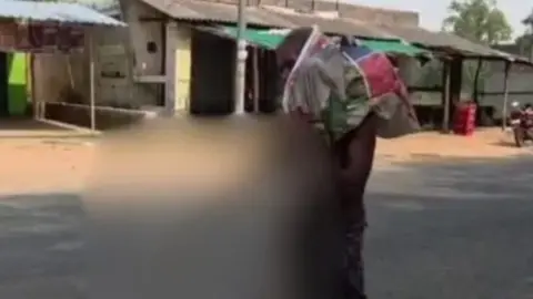 A man wearing an Indian dhoti - which is a piece of cloth wrapped around the waist - is seen carrying a sack-like object over his shoulder. These are the skeletal remains of his sister which have been blurred. 