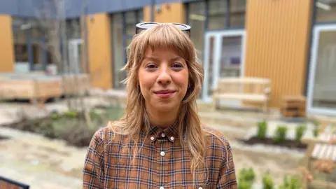 Katy is standing in the courtyard of Shankill Shared Women's Centre. She has blond wavy hair with a fringe and has brown sunglasses on top of her head. She is wearing a dark brown checkered shirt. She is smiling at the camera. 