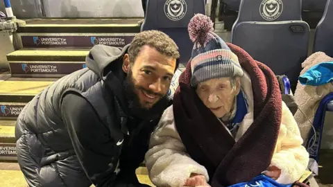 Cosham Court Nursing Home Joan, an elderly woman in a hat and with a blanket around her shoulders, is seated while a bearded man bends into shot and smiles in the direction of the camera. 