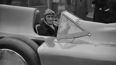 A black‑and‑white photo of a person in a vintage open race car, wearing a cap and goggles, with a few onlookers in the background.
