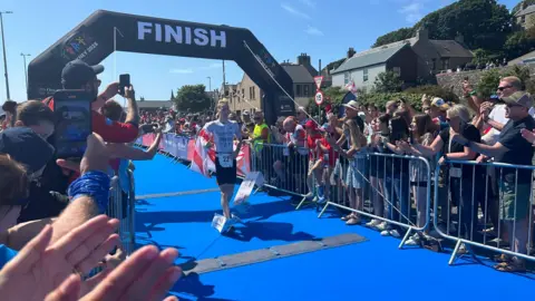 BBC A runner crosses a blue-floored finish line beneath a large “FINISH” arch while spectators line both sides of the course, cheering and taking photos on a sunny day.