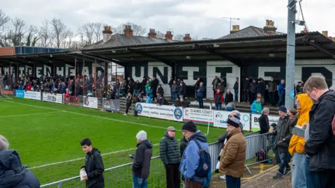 Fans wearing hats and coats are gathered around the pitch at a small football ground. Across the far wall, the words 'Maidenhead United' are spelled out in large letters across a wall. 