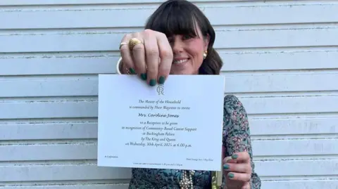 Caroline Jones Caroline Jones stands against a white panelled background. She wears a green floral dress and is holding her invitation which is printed on white card. She has brown shoulder length hair with a fringe and wears pearl earrings.