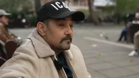 Stephen Hart in a cream jacket and black baseball cap sitting on a park bench.