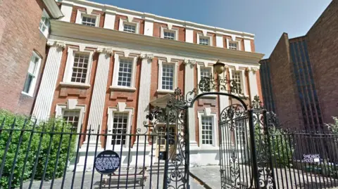Google Number 11 Priory Row, Coventry. The three-storey building has rectangular windows and in the front are green bushes and black railings as well as an open gate which has a curved top. 