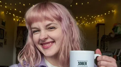 A young woman with pink hair in a bedroom with fairy lights on the ceiling. She is wearing a purple top and is holding a white mug that says "sober" in black writing