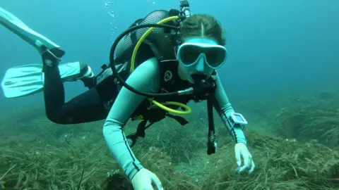 Mia is seen scuba diving near the seafloor, wearing a mask, fins, and full diving gear. Bubbles rise from her regulator as she glides over rocks and sea grass in clear blue water.