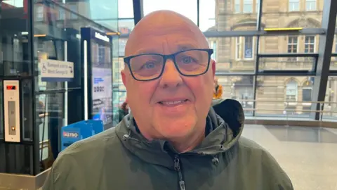 Tommy Whitelaw, who has a bald head and is wearing glasses, is seen from the mid-chest up. He appears to be middle-aged, has a friendly expression and is smiling. He is wearing a dark olive green or greyish-green jacket or windbreaker with a zipper and a hood. In the background is a train station with large windows with reflective glass showing a city street scene beyond, including a light-coloured building with arched windows.