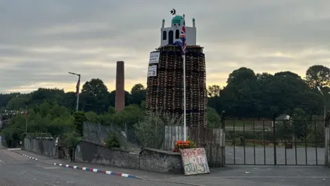 An image of the top of the bonfire, consisting of wood pallets, with a structure resembling a mosque on top. Two placards are below. One reads "secure our borders" while another reads "end the threat of radical Islam".