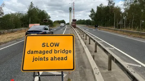 BBC/Richard Edwards This image, at the entry to a river bridge has a yellow and black sign warning that the bridge's joints are damaged and traffic must drive slowly. A red-painted sign further ahead warns drivers they will cross a ramp. Traffic is heading along one side of the bridge, away from the camera.