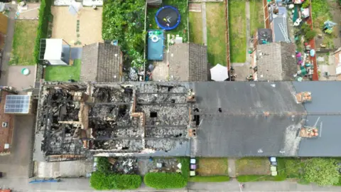 A drone shot of the burnt out houses from above.  Three of the houses are completely burnt out and there are several holes in the fourth to the right.