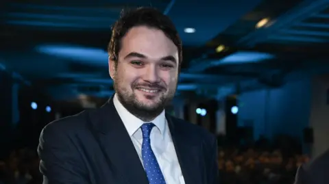 Reform UK Robin Hunter-Clarke is wearing a dark suit and blue spotted tie. He is inside, in a dimly lit room.