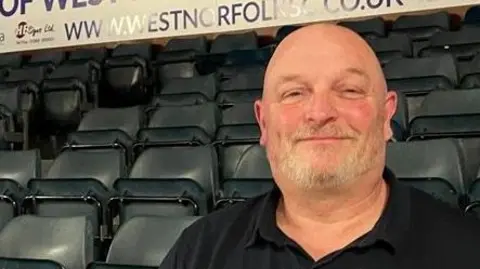 Clare Worden/BBC Simon Rose chair of West Norfolk Swimming Club sits in a bank of grey seats in the spectators' area of St James Swimming Pool. He is bald, has a grey beard and wears a black t-shirt. He smiles at the camera. 