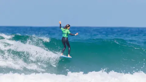 Jason Feast A person wearing a green top and black wetsuit balances on a surfboard while riding along the crest of a small ocean wave. White foam splashes behind the surfboard, with deep blue water and a clear sky stretching across the background.