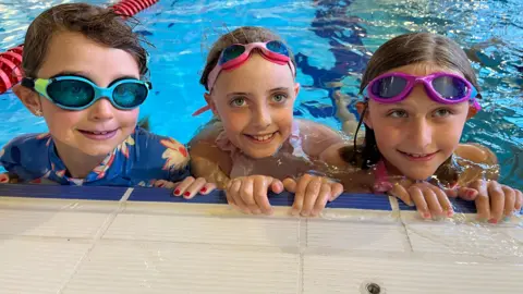 BBC/Lucinda Adam Three girls aged 7 and 8 look up from the side of a swimming pool. They are submerged to their shoulders and all three have goggles on their heads.