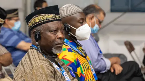 Ahmadiyya Muslim Jamaat Men from different ethnic backgrounds are seated next to each other at an indoor seminar. Two of them wear headphones.