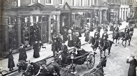 Bath Record Office Archive photo of a parade on Bath's Milsom Street (outside Jolly's department store) in 1908. The black and white image shows people in military uniform and Victorian clothes walking on the street and horses, some pulling carts, walking down the street.