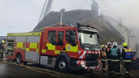 Fire engine positioned in front of the damaged cottage. Four fire fighters are positioned to the right of the vehicle. A sixth fire fighter is stood at the rear of the vehicle to the left. The cottage appears damaged on its rights side by the chimney.