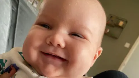 Contributed A baby boy smiles at the camera in a close up picture. He is wearing a white baby grow while an adult holds him.