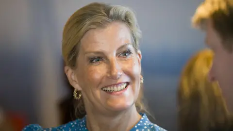 PA Media The Duchess of Edinburgh is smiling at someone off camera. She has blonde hair tied back from her face and blue eyes. She is wearing ornate gold hoop earrings and a blue and white spotted top.