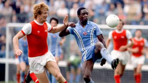 Getty Images Two men are playing football and wearing football kits while standing on a football pitch.