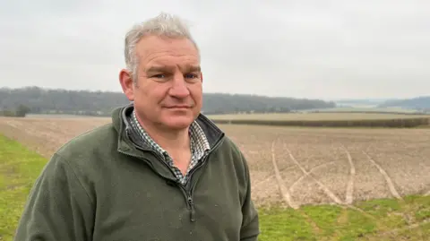 In the foreground the farmer David Lemon stands in a green fleece, in the background large scars can be seen on the field, from the tyres of hare courser's vehicles.