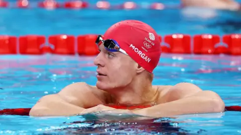 Maddie Meyer/Getty Images Oliver Morgan is in a blue swimming pool with red lane dividers. He is wearing blue swimming goggles on his forehead and a red swimming cap