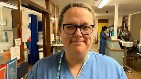 A lady wearing nurse's scrubs, with tied back brown hair and glasses smiles at the camera in a hospital ward, with people working at a desk over her shoulder. She is wearing an NHS lanyard. 