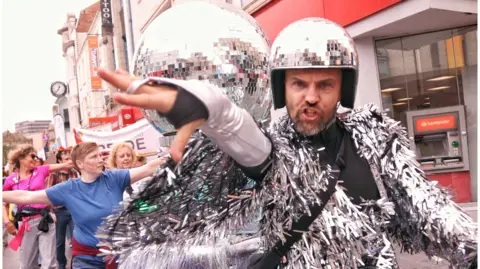 Andi Talbot A man wearing a silver tinsel jacket and a helmet which looks like a disco ball. He is staring into the camera with his hand held out towards it. He is mid-speech with his mouth open and he has a grey beard. People behind him are dancing down Northumberland Street.