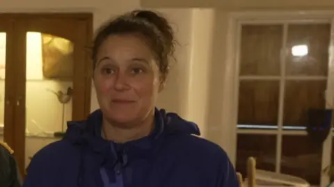 The image shows a person standing indoors wearing a dark blue hooded jacket. Behind them is a wooden display cabinet with glass doors containing various household items. To the right is a window with multiple panes.