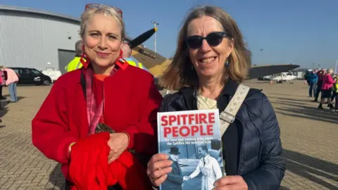 Two women smile at the camera. A blonde woman wearing a red coat and brunette with sunglasses is holding a book called Spitfire People, she is wearing a blue jacket. They are standing in front of a Spitfire and an aircraft hanger