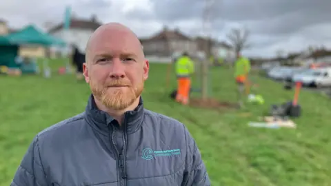 A man stands in a green space in Whitleigh, Plymouth. There are people in high-viz clothing planting trees in the background. You can see semi-detached houses around the end of the green space. 
