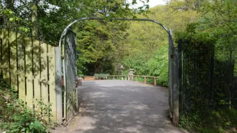 Metal gates at one of the entrances to Saltburn's Valley Gardens. They are open, leading on to a path with trees in the background. Wooden fences stand either side of the gates.