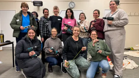 Sarah Bolger and Justine Wortsman taking a group photo with participants on one of their taster sessions. All are smiling. Sarah has three silver necklaces around her neck and is wearing a black shirt. She has long, brown hair.