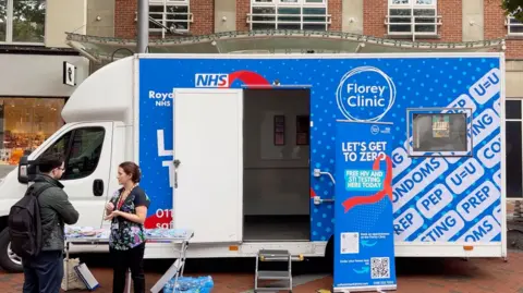 Blue coloured van that has a sexual health sticker a banner that says let's get to zero and Kate Rabjohns speaking to a man. They are standing in front of a table with different leaflets. 