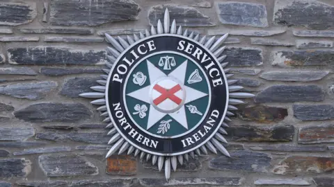 A silver, green and black circle sits on a grey-stone wall. The badge has the words 'Police Service Northern Ireland' around its circumference. 