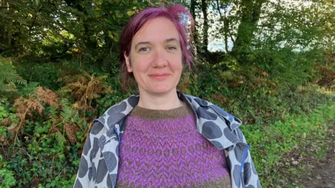 Una Devlin from the NHS Forest project is wearing a purple jumper with a blue coat standing in a green space at Langdon Hospital, Dawlish.