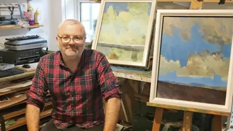 Paul Bailey Paul Bailey sits in an art studio surrounded by pieces of his work. He has short grey hair and wears a red and black chequered shirt. He wears glasses and rests his hands on his lap. 