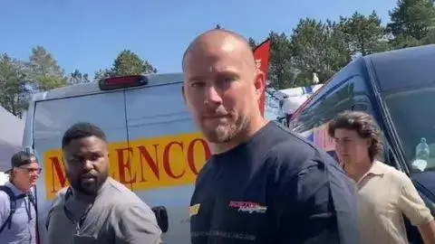 Channing Tatum a bald man looks and smiles he wears a navy t-shirt and stands in the paddock with vans in the background.