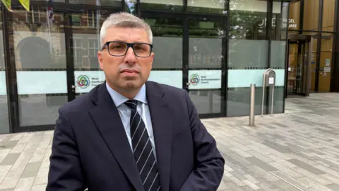 Ben Schofield/BBC A man standing in front of a glass-fronted building, wearing a blue shirt, a tie and a blue jacket. He is standing on paving slabs. He has short grey hair and is wearing glasses.