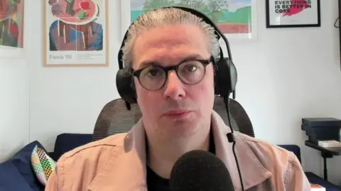 Kevin McPartlan is on an online chat. He's wearing headphones and is sitting in front of a mic. He's in a living room and is wearing a salmon-coloured overshirt. Behind him on the white wall are various works of art.