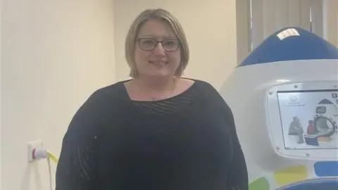 MFT Charity Fundraising Manager Terri Torevell stood in front of the device. She has short blonde hair and is wearing glasses with a black jumper.